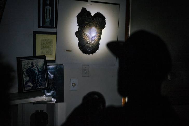 In a dark room, a flashlight illuminates an African mask that hangs on a wall.
